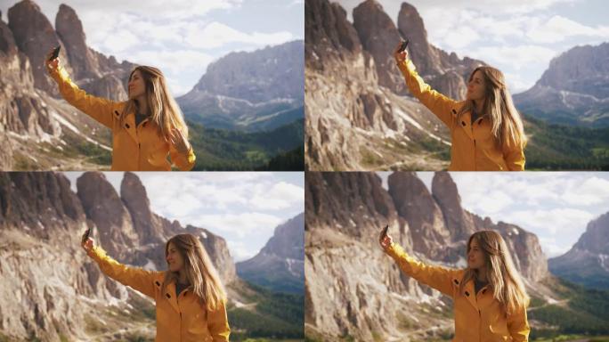 穿着黄色雨衣的年轻开朗的女人在阳光明媚的日子里在美丽的山脉背景上自拍，慢动作