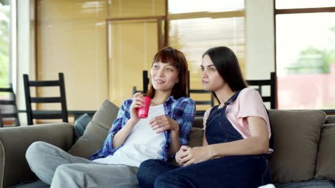 平移两个不同的女性朋友，亚洲和高加索人，坐在家里的沙发上，看连续剧，电视节目或电影并讨论它。女人说话