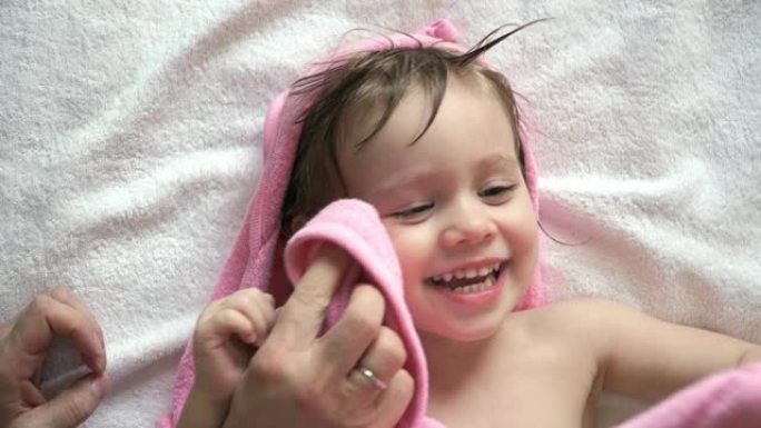 洗澡后，穿着带兜帽的粉红色毛巾的小女孩的肖像躺在床上玩耍和大笑。父亲的手擦着微笑女儿的湿脸和头发。