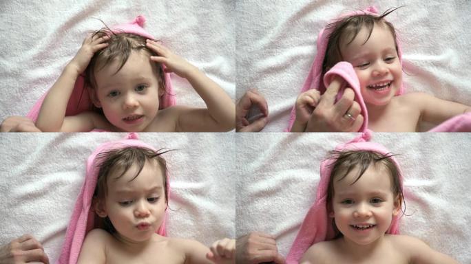 洗澡后，穿着带兜帽的粉红色毛巾的小女孩的肖像躺在床上玩耍和大笑。父亲的手擦着微笑女儿的湿脸和头发。
