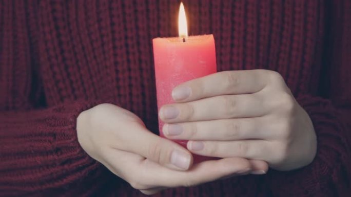 在红色舒适的汗衫背景上，女性双手燃烧红色蜡烛。年轻女子拿着燃烧的蜡烛。用蜡烛祈祷。乞讨概念。