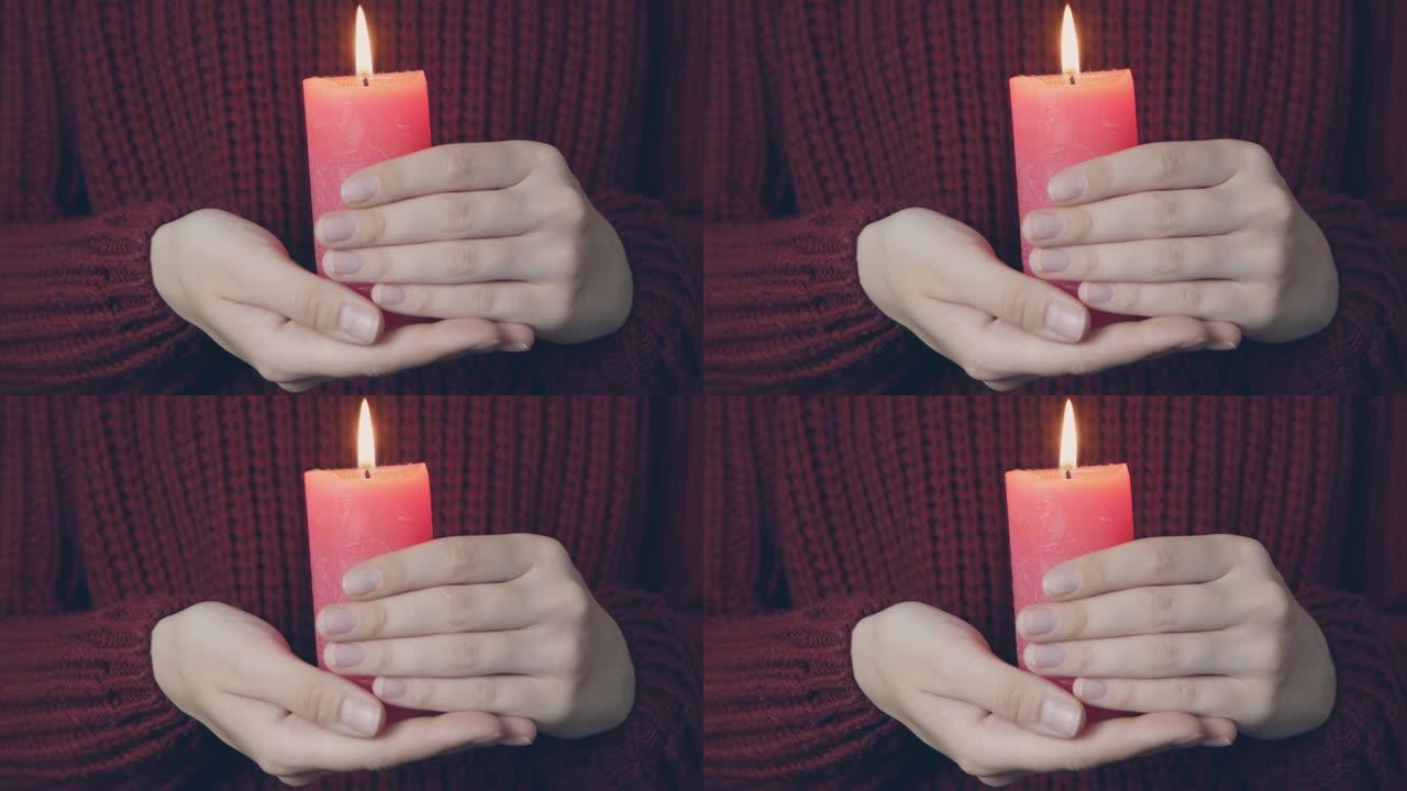 在红色舒适的汗衫背景上，女性双手燃烧红色蜡烛。年轻女子拿着燃烧的蜡烛。用蜡烛祈祷。乞讨概念。