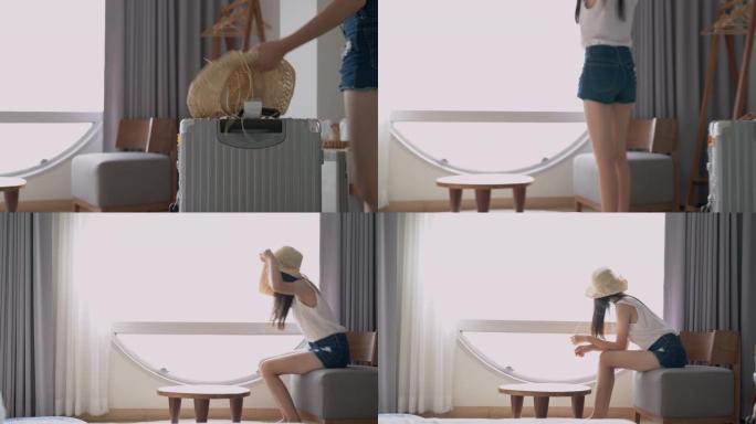 年轻美丽的亚洲女人在手提箱上摘帽子，坐在沙发上舒适舒适地靠近窗户，为夏天在酒店房间的下一次旅行做好准