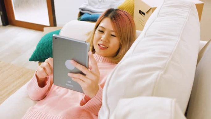 亚洲妇女躺在沙发上，在家中使用平板电脑，情绪低落。数字时代的生活方式。