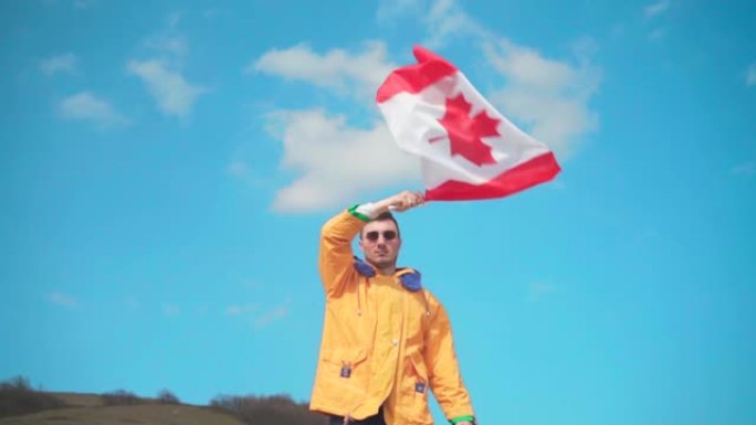 一个穿着黄色夹克，蓝色牛仔裤和眼镜的男人站在山上，从一侧到另一侧挥舞着加拿大国旗。