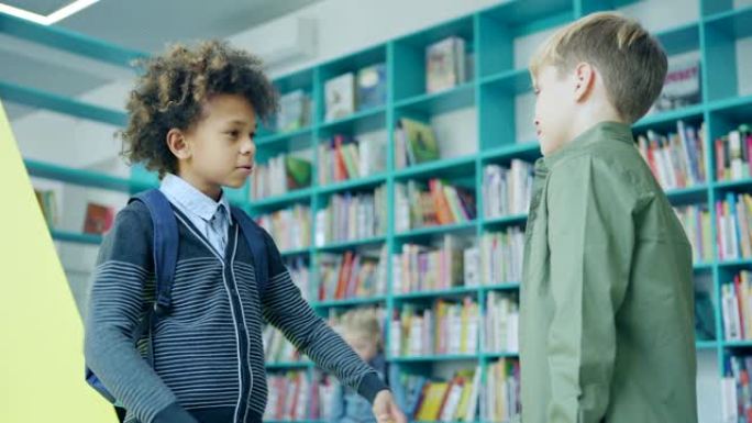 两个男小学生朋友，混血和高加索男孩，在学校图书馆的书架旁站着友好的谈话