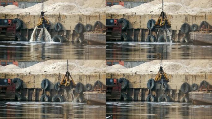 挖掘机的抓斗从河中升起，港口的水溅起，背景是旧的，生锈的老式驳船和汽车轮胎