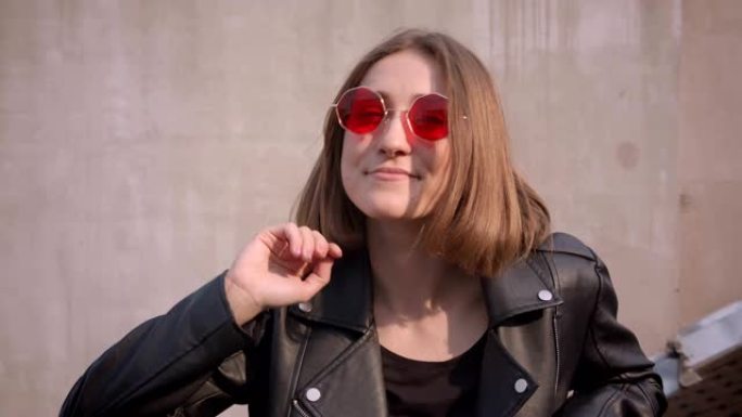 穿着皮夹克和红色时髦太阳镜的年轻可爱的高加索女孩的特写肖像在城市户外快乐地看着相机