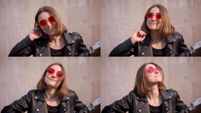 穿着皮夹克和红色时髦太阳镜的年轻可爱的高加索女孩的特写肖像在城市户外快乐地看着相机