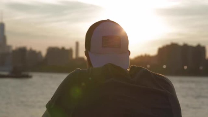 白人男子戴着白色帽子从布鲁克林俯瞰纽约市。美丽的太阳光。水和船。下午结束时的日落。