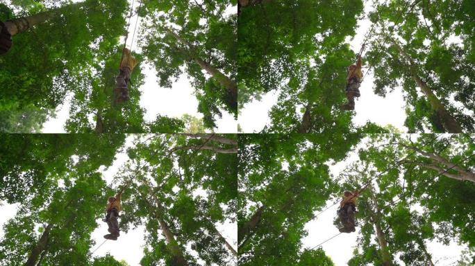 在森林冒险公园的树梢上，一个穿着安全带的小男孩的慢镜头在一条路线上攀爬。他爬上高高的绳索小径。户外娱