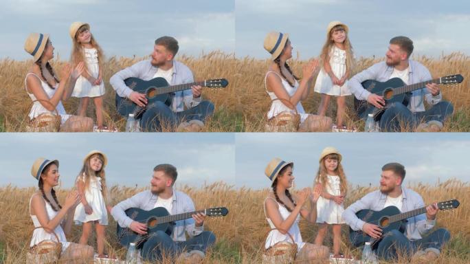 家庭户外娱乐，年轻的男子弹吉他，而他的女人和可爱的小女儿在草帽和白色的衣服拍手在麦田在收获的时候露天