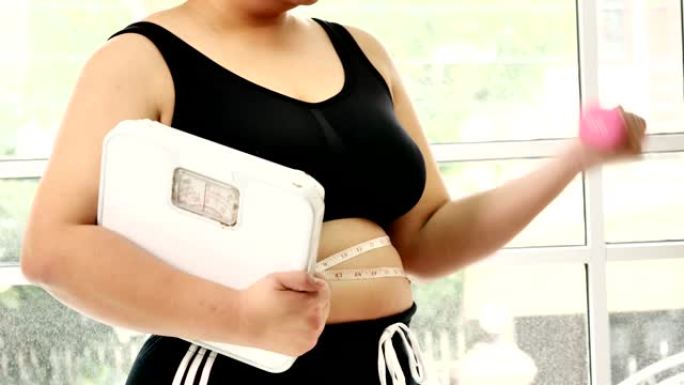 穿着运动服的大身材年轻女子，手持体重秤和哑铃