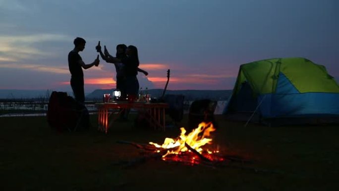 剪影一群亚洲朋友游客在日落时在湖边露营时，夏天喝酒精啤酒和弹吉他与幸福