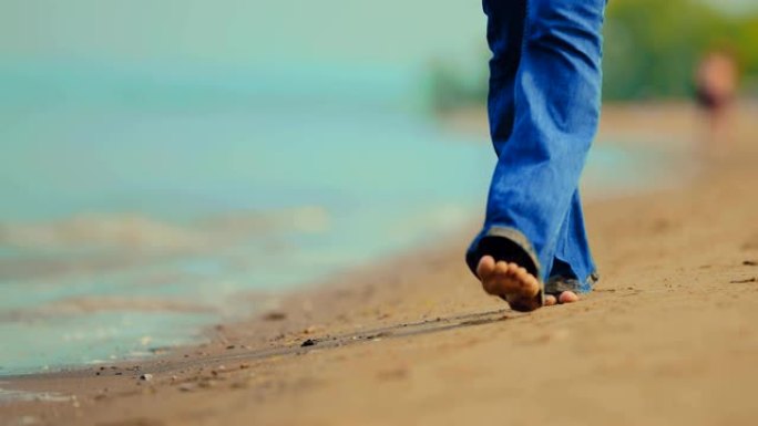 穿着湿牛仔裤和赤脚的女人走在沙滩上的沙滩上。沿着海边散步。特写。