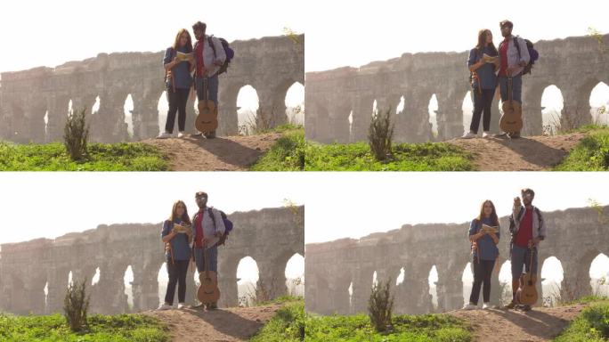 年轻可爱的夫妇背包客游客阅读地图指向罗马渡槽拱门在罗马公园废墟上浪漫的迷雾日出与吉他