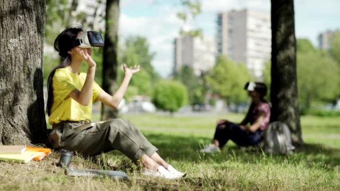 亚洲女学生在VR耳机中滑动可视化数据坐在公园的树下，在她的膝盖上的教科书。另一个在背景中戴着虚拟现实