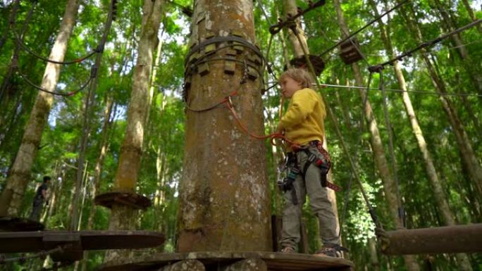 在森林冒险公园的树梢上，一个穿着安全带的小男孩的慢镜头在一条路线上攀爬。他爬上高高的绳索小径。户外娱