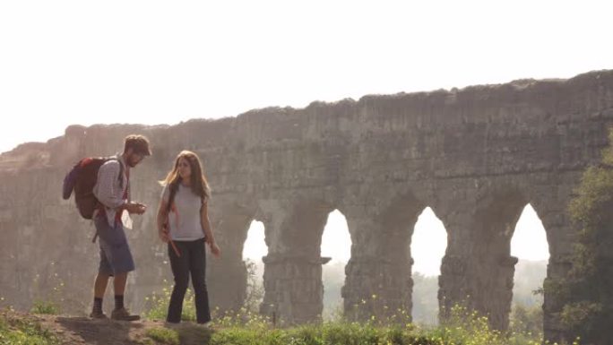 年轻可爱的夫妇背包客游客阅读地图指向罗马渡槽拱门在罗马公园废墟上浪漫的迷雾日出与吉他