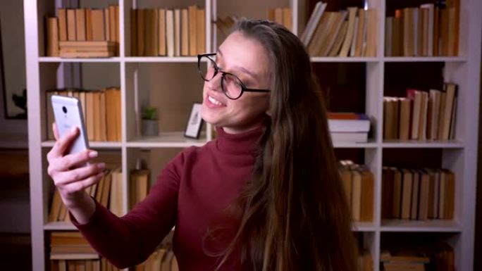 戴着眼镜的年轻高加索女学生的特写肖像在室内的大学图书馆里，在电话上进行视频通话