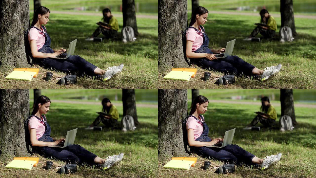 女学生坐在树下的侧视图，靠在树干上，在公园的笔记本电脑上打字; 附近有教科书，咖啡杯和VR眼镜。背景