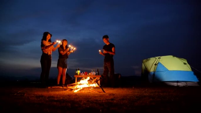 场景慢动作青少年亚洲人在暑假期间在帐篷前与篝火一起享受，并在湖边玩烟花。他们在假期，放松的时刻快乐并
