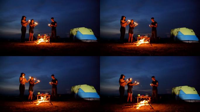 场景慢动作青少年亚洲人在暑假期间在帐篷前与篝火一起享受，并在湖边玩烟花。他们在假期，放松的时刻快乐并