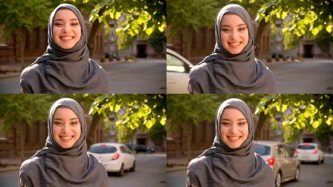 戴着头巾的穆斯林学生的肖像站在绿色城市街道上快乐地对着镜头微笑。