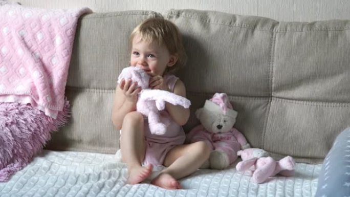 小1岁女婴的肖像坐在沙发上，带着毛绒玩具，微笑着玩耍。希尔德拥抱泰迪熊玩具并亲吻它。