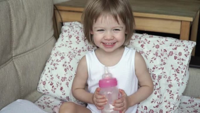 小可爱的女婴的特写镜头早上醒来坐在床上，手里拿着一瓶牛奶，微笑着笑。
