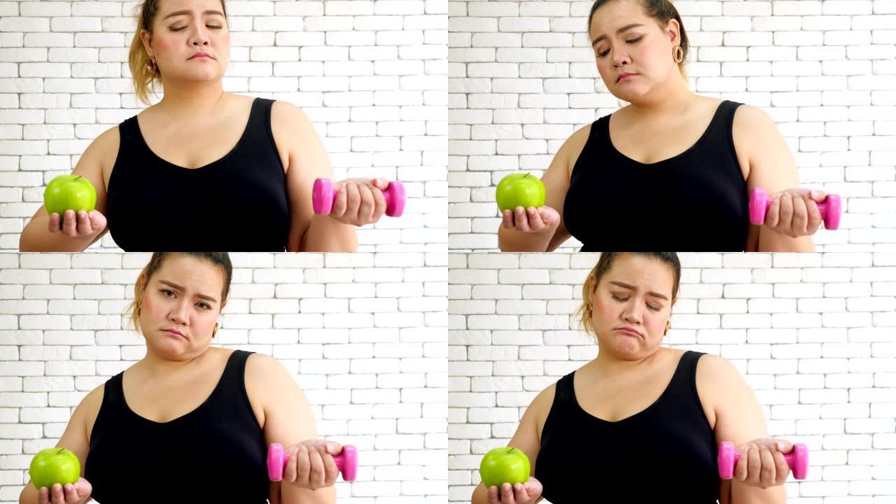 穿着运动装的不快乐的超重女人的肖像拿着哑铃和苹果水果