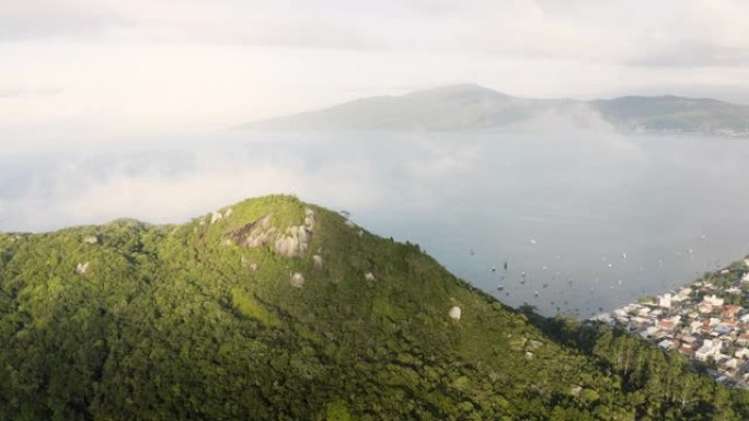 在一个夏日，无人机在一个热带森林山的云峰周围拍摄，背景是城市和海洋