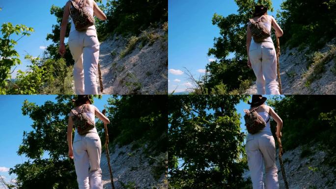 一个背着背包和木棍的女孩旅行者正沿着一条位于山中陡坡上的小路行走。