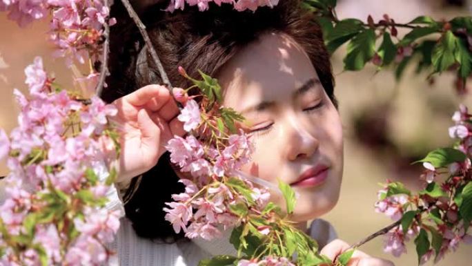 穿着白色衣服的美丽中国年轻女孩在微风中闭上眼睛，享受阳光明媚的春天，粉红色的樱花森林背景，4k电影，