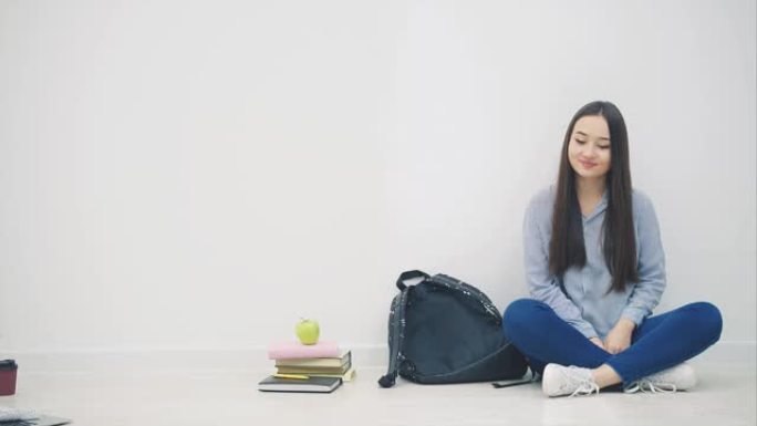 可爱的年轻亚洲女孩出现在白色背景上，以lotos的姿势坐在地板上，挥手打招呼，打开背包，拿出素描本。