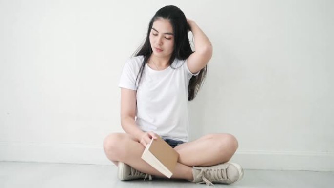 亚洲少女拿着棕色的书，坐在地板上摸着头发，慢动作。