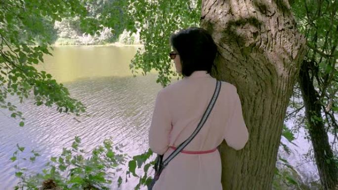 中年妇女靠在池塘旁，靠在树上