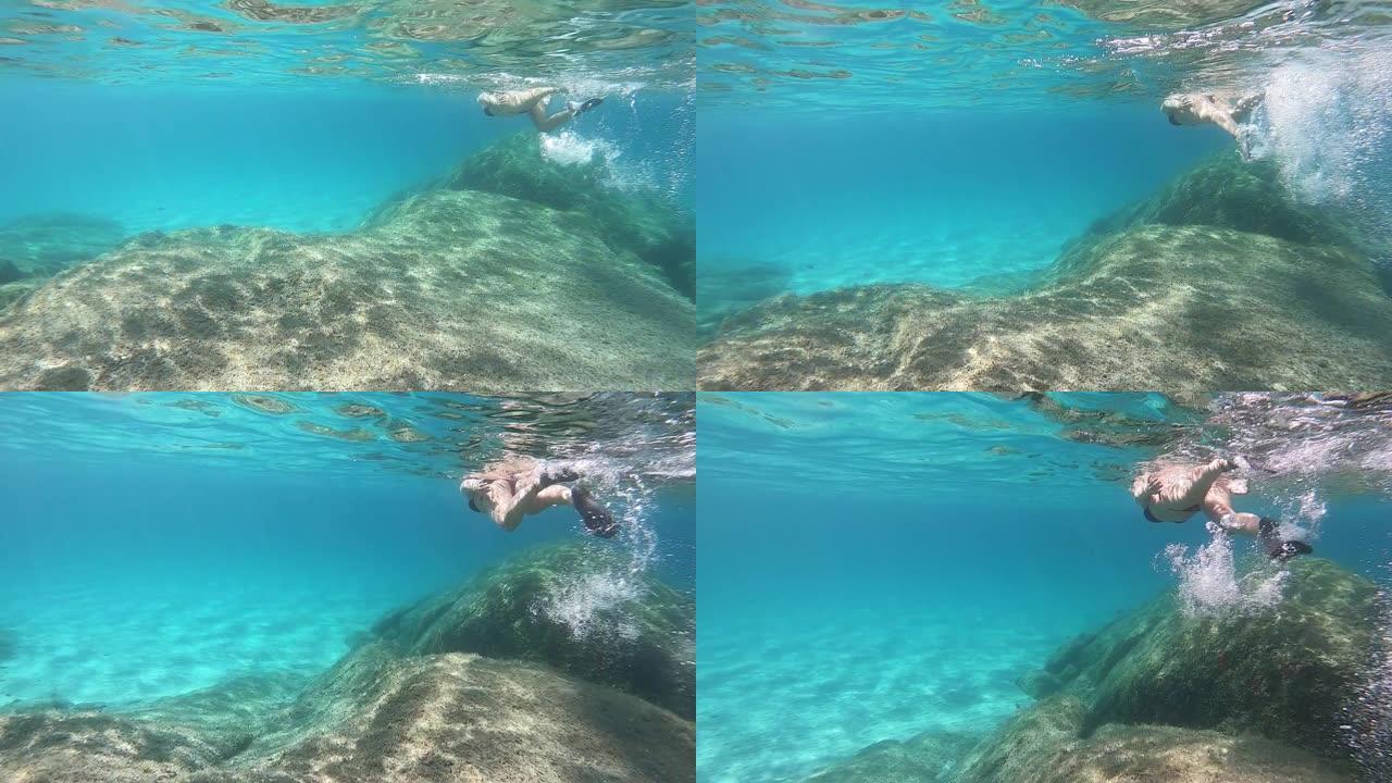 一个穿着黑色比基尼的美丽年轻女子的慢动作从水下游泳在蓝色的水中戴着面具，在岩石和彩色海洋植被和白色沙