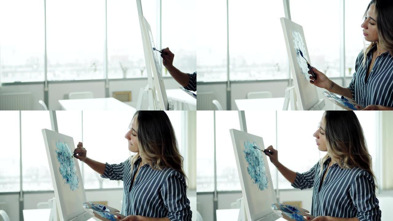 在艺术治疗课上跟踪站在画架旁用调色板刀和蓝色丙烯酸颜料创作抽象画的美丽年轻女子的右侧视图