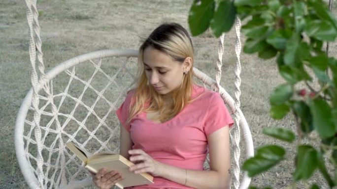 一个年轻的女孩，金发碧眼，在阳光明媚的日子里，在公园的白色秋千上看书，在户外，在树荫下。她微笑着翻开