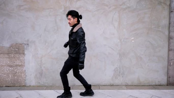 中国年轻的胡子男人在城市混凝土墙旁边的街道上跳舞。他穿着夹克，看起来有点尴尬，4k镜头，慢动作。