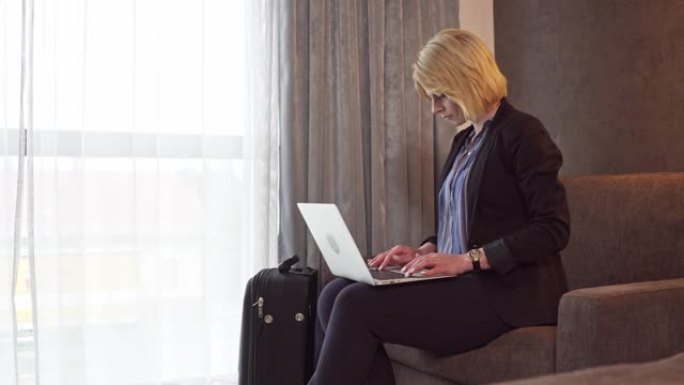 穿着黑色西装的金发女商人在酒店房间里用笔记本电脑工作