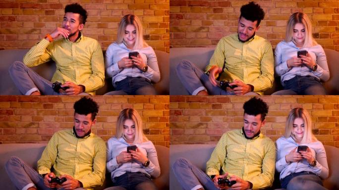 非洲家伙用心用操纵杆玩电子游戏，他漂亮的高加索女友在舒适的家里看着手机。