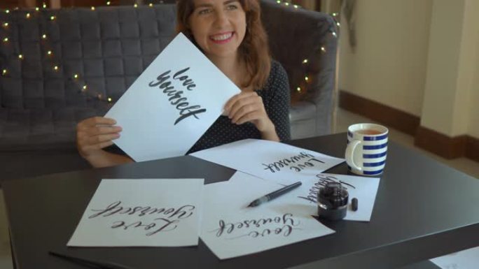 一个年轻女子使用刻字技术在纸上写字的特写镜头。她看着床单，上面写着爱自己的文字，笑着