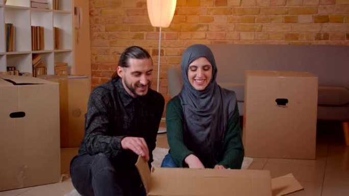 年轻幸福的穆斯林夫妇的特写镜头坐在新买的公寓的地板上，愉快地微笑着，建议在哪里挂照片