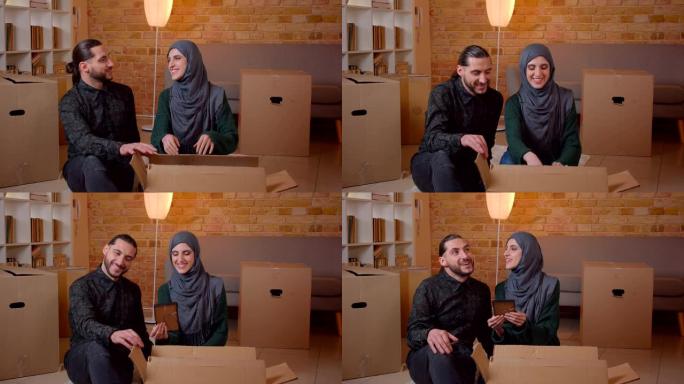 年轻幸福的穆斯林夫妇的特写镜头坐在新买的公寓的地板上，愉快地微笑着，建议在哪里挂照片