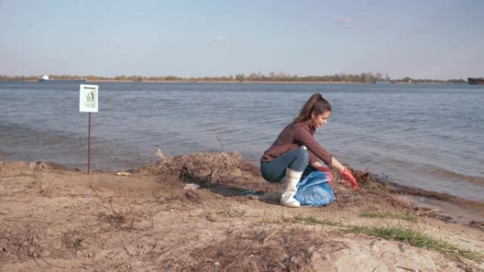环境解决方案，女孩志愿者在垃圾袋中收集塑料垃圾，并在靠近水的受污染的河滩上做出积极的姿态