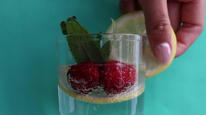 桌子上是一杯矿泉水。在水中放一片柠檬，草莓和新鲜的薄荷叶。可见美丽的气泡游戏。一个女人把柠檬片放在玻