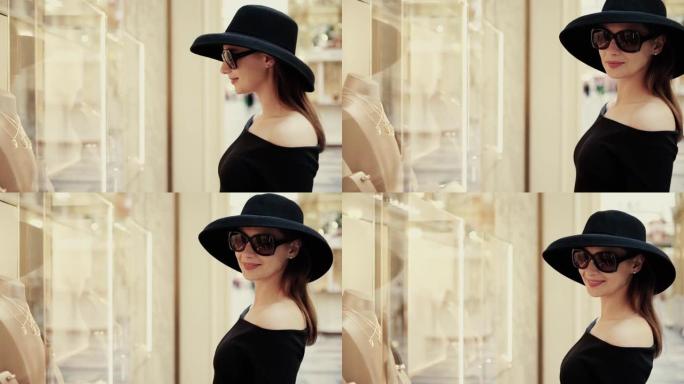 一个戴着帽子和太阳镜的漂亮时尚女孩有兴趣看精品店的橱窗