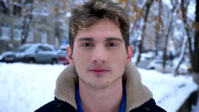在下雪的日子里，穿着冬衣站在户外的年轻迷人的白人男性的特写镜头直视相机